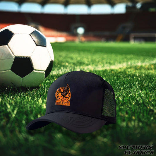A black trucker-style baseball cap with a curved brim and mesh back panels, featuring an orange embroidered eagle logo and the word 'MEXICO' on the front. The cap is placed on a green grass field with a soccer ball in the background, with a blurred stadium visible behind it.