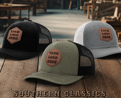 Three trucker-style caps displayed on a wooden table in a rustic shop setting: one black, one light gray, and one sage green. Each cap has a tan patch on the front with placeholder text reading 'YOUR LOGO HERE.