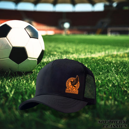 A black trucker-style baseball cap with an orange Mexico soccer emblem on the front, placed on a green grass field with a soccer ball in the background.