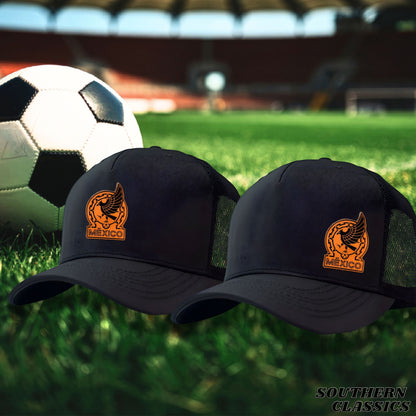 Two black trucker-style caps with an orange Mexico soccer emblem on the front, displayed on a green grass field with a soccer ball and stadium in the blurred background.
